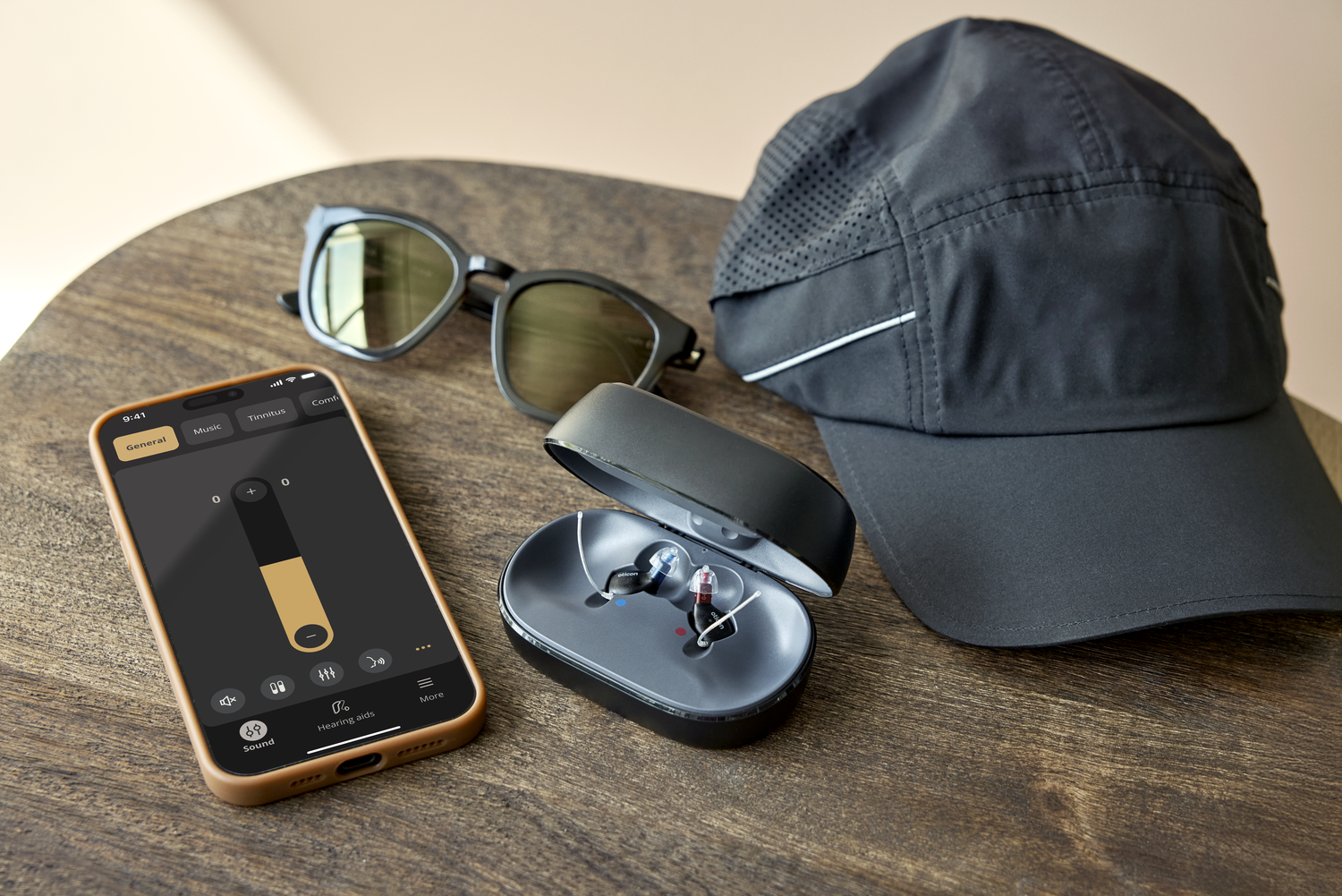 Oticon Zeal hearing aids in charger with smartphone app, sunglasses, and hat on a table
